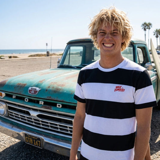 Person standing next to a vintage Ford truck with a surfboard, wearing a stripe black and white Wax Zombies t-shirt.